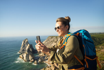 Happy female backpacker taking selfie at precipice. Mid adult woman wearing sunglasses and backpack...