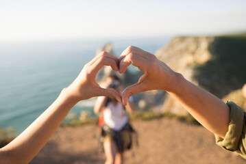 Close-up of female hands shaping heart outdoors, teenage girl standing in background. Female family...