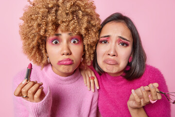 Upset shocked disappointed mixed race women look sadly at camera hold lipstick and eyelashes curler apply makeup undergo beauty procedures isolated over pink background attend makeup courses