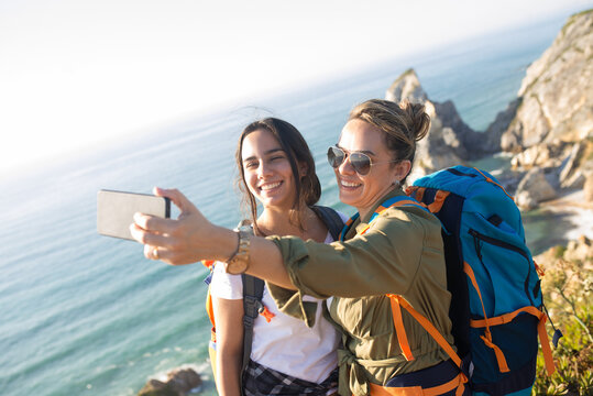 Portrait of mother and daughter taking selfie with seascape. Happy mid adult woman hiking with teen girl in summer. Active family weekend concept