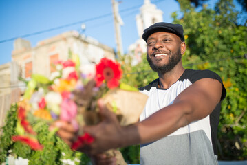 Close-up of happy African American man presenting flowers. Smiling young man in casual clothes standing looking at partner giving bouquet of beautiful flowers. Dating, romance, relationships concept