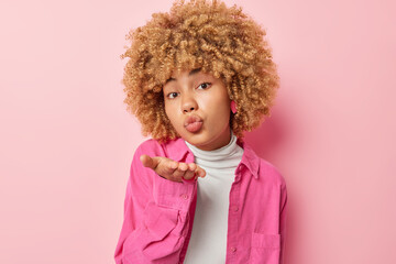 Cute romantic European woman blows air kiss sends mwah at camera has tender facial expression keeps lips rounded wears white comfortable turtleneck and shirt isolated over pink background adores you