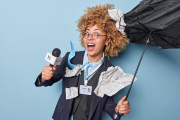 Happy positive curly haired female news reporter holds microphone exclaims positively dressed in...