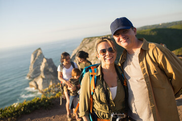 Fototapeta premium Happy couple of backpackers hiking with children. Mid adult man and woman posing with their family in background. Active family weekend concept