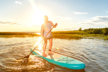 a man in shorts and a T-shirt on a SUP board with an oar floats on the water against the background of the sunset.
