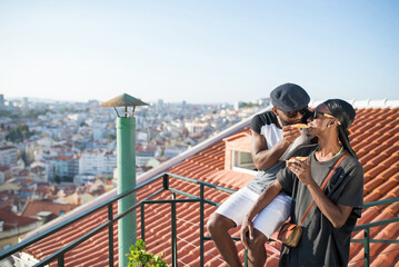 Portrait of happy gay couple dating with tasty pizza. Two bearded African American men feeling joy high on roof top hugging feeding each other slices of pizza. Same sex couples love, happiness concept