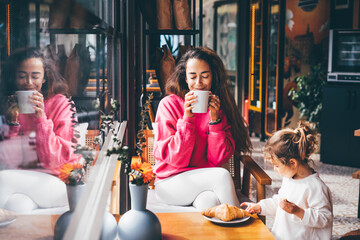Happy woman with daughter drinking coffee with croissant in cafe.