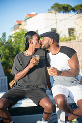 Portrait of happy African gay couple kissing on bench. Two men in casual clothes sitting on bench with glasses of juice kissing each others lips. Homosexual love and relations of LGBT couples concept