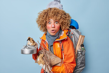 Photo of scared curly haired young woman carries wood and kettle carries heavy backpack wears warm...