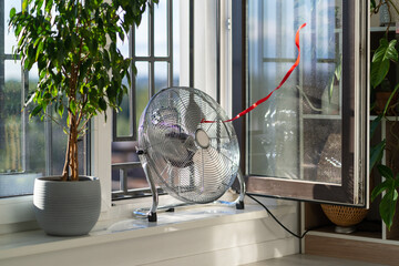 Working electric fan with red wriggle ribbon designed to cool air and supply wind is located on windowsill next to house plant. Equipment for cooling temperature in room in hot summer weather