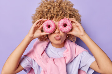Photo of millennial girl with curly hair covers eyes with delicious donuts keeps lips folded enjoys...