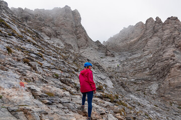 Naklejka premium Woman climbing on mystical foggy hiking trail leading to Mount Olympus (Mytikas, Skala, Stefani) in Mt Olympus National Park, Thessaly, Greece, Europe. Scenic view of cloud covered slopes and ridges
