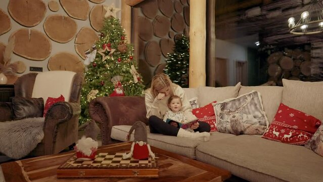 Full shot - A young mom with her baby on her lap, sitting on a beige and dark brown sofa, with Christmas pillows and a Christmas tree behind them, holding a book and interacting with her baby, on a Ch