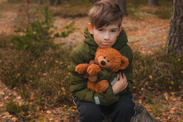 Child alone in the forest with a teddy bear. A boy in solitude in an autumn forest