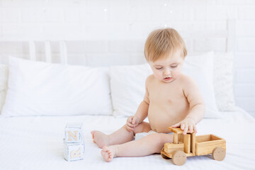 cute blonde baby boy is sitting on a white bed at home in diapers and playing with a wooden toy car