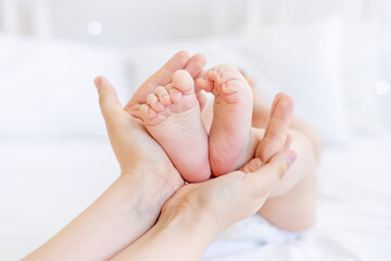 Obraz na płótnie Canvas mom's hands hold the baby's feet in focus on a white crib at home with white cotton bedding, the concept of baby goods and accessories and mom's love and care