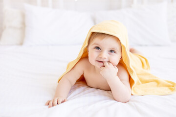 Fototapeta na wymiar cute blonde baby boy with a yellow towel on his head after a bath or bathing on a white bed at home in a bright room smiles, the concept of child care and hygiene