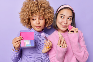 Two mixed race women apply daily makeup hold palette and concealer take care of appearance want to look beautiful dressed in casual clothes isolated over purple background enjoy new makeup cosmetics