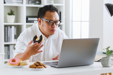 Asian fat man eating donut, sweet, junk food during working with computer laptop, unhealthy eating...