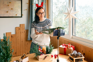 Female florist blogger making winter ikebana with fresh pine branches, candle and christmas decorations in streaming in front of her smartphone. Small business. Seasonal winter workshop.
