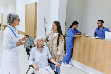 Fototapeta premium Patient care in hospital. Experienced doctor talking and advising with relative woman of patient and take care of health of senior woman sitting on wheelchair