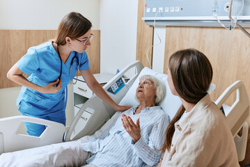 Nurse caring for elderly patient lying in bed of hospital ward, elder care. Daughter supports elderly mother patient during round of ward nurse