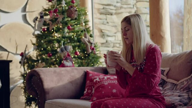 Medium shot - A blonde woman having a cold and sneezing, then sipping from a mug, while sitting on a couch with Christmas print pillows and a Christmas tree in the background, in daylight.