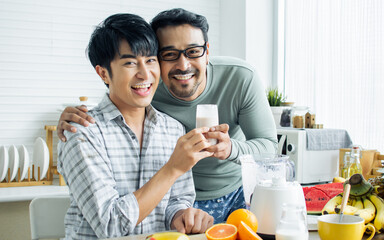Gay LGBT sweet Asian couple wearing pajamas, smiling, with happiness and love while making and pouring juice smoothie to glass and serve to lover in kitchen at home in morning. Lifestyle Concept.