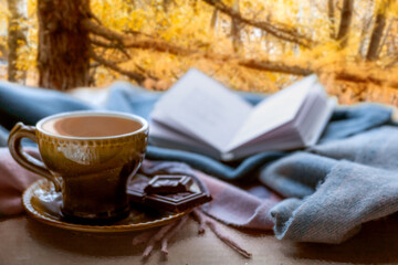 The idea of coziness, hugge, home comfort. Autumn light sadness. Coffee, chocolate, a blanket and a book on the background of a forest landscape on an autumn evening