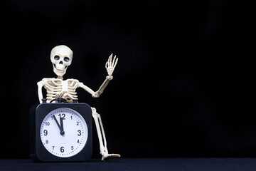 A human skeleton waving his hand, greeting holding an black alarm clock on a black background, copy...