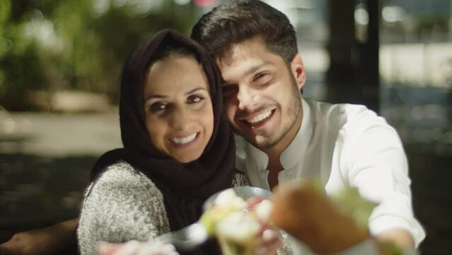 Happy muslim couple showing their lunch to camera on sunny day. Young Arabic man and woman smiling, spending time together outside. Front view camera. Joy, friendship, romance concept. 