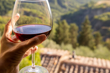 Woman holding a glass of red wine with beautiful landscape of Italy in a background on a sunny day. View from the window.
