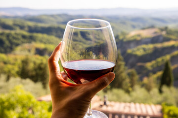 Woman holding a glass of red wine with beautiful landscape of Italy in a background on a sunny day. View from the window.