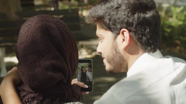 Close-up of young muslim couple doing selfie outside. Woman in hijab taking photo pressing shutter button on mobile phone while dark-haired man embracing her. Modern technology, relations concept. 