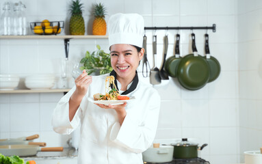Portrait of Asian young beautiful chef woman wear white chef uniform and hat, holding spaghetti dish, smiling looking at camera with delicious face, working at restaurant kitchen