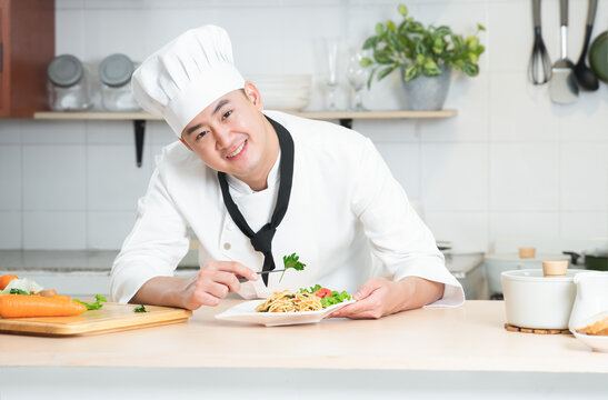 Portrait of Asian young handsome chef man wear white chef uniform and hat, smiling looking at camera, holding food forceps to garnish spaghetti dish, working at restaurant kitchen