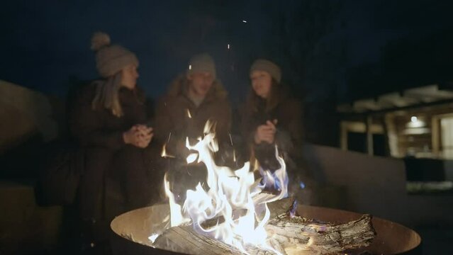 Bokeh - Friends gathered around the outdoor fire pit at winter night. Three people in winter attire, talking and hanging out on a calm winter evening