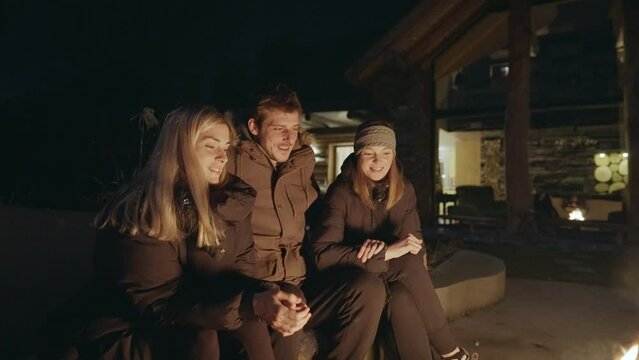 Medium shot - A group of three, a man and two females, hugging while sitting next to a fire, with a cottage in the background, dressed in warm jackets, happy on a calm night.