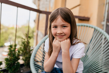 happy adorable little girl with lovely smile wearing white t-shirt sitting on the balcony in warm sunny summer day
