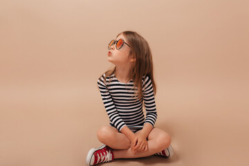 Stylish happy little girl inn striped dress and stylish glasses sitting one the floor and looking...