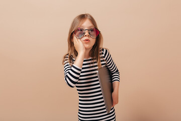 Incredible little girl in striped dress and stylish glasses touching her cheek and looking aside with laptop over beige background 