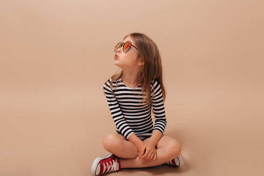 Stylish happy little girl inn striped dress and stylish glasses sitting one the floor and looking aside over isolated beige background 