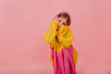 Photo of little girl with happy smile in bright raincoat covering face and smiling while looking into camera on pink background. 