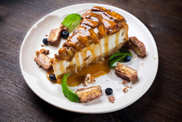Caramel-flavored cheesecake. A slice of dessert topped with salted caramel syrup, peanuts and milk chocolate. The cheesecake is garnished with blueberries and mint leaves.