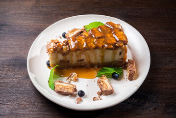 Caramel-flavored cheesecake. A slice of dessert topped with salted caramel syrup, peanuts and milk chocolate. The cheesecake is garnished with blueberries and mint leaves.