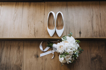 The bride's bouquet with a ribbon and white shoes lie on a wooden floor. Wedding photography, top view.