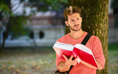Fototapeta premium Young male student reading a book lying against a tree in the park
