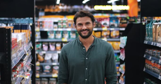Attractive multi-ethnic dark-haired guy in casual wear standing with a shopping cart in the supermarket while making purchases, buying goods, smiling a beautiful toothy smile looking at the camera