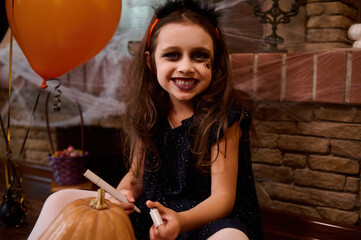 Mischievous child, a little girl dressed as a witch with a spider painted on her face, smiles looking at camera, drawing a scary face on a pumpkin, a Jack-O-Lantern. Concept of a happy Halloween party