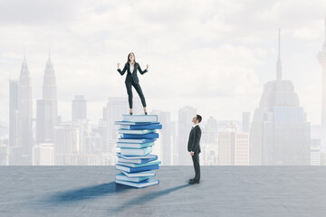Young european businessman looking up at happy woman standing on book pile. Blurry city background. Education, knowledge and success concept.